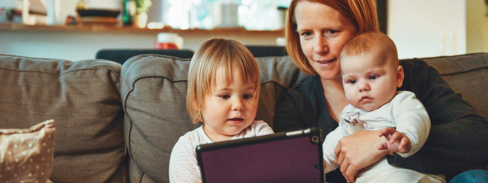 photo-1476703993599-0035a21b17a9 Mom holding her baby while sitting next to her toddler and showing them both a tablet.